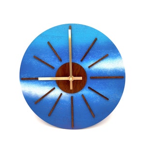 May include: A blue and white wooden clock with gold hands and a brown center. The clock has a sunburst design with wooden rays.