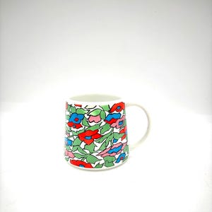 May include: White ceramic mug with a colorful floral pattern. The design features red, blue, and pink flowers with green leaves. The mug has a simple, curved handle and a clean, white interior.