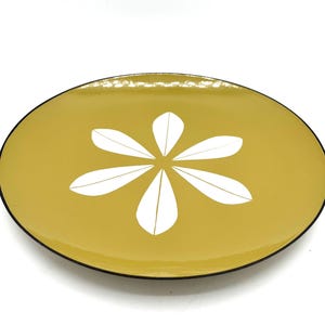 Pode incluir: Prato ovalado amarelo mostarda com um desenho floral branco no centro. O prato tem uma borda preta. O desenho é uma flor estilizada com seis pétalas brancas. O prato é provavelmente feito de cerâmica ou esmalte.