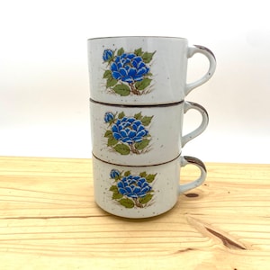 May include: A stack of three white ceramic mugs with blue floral designs. The mugs have brown speckled glaze and brown handles.