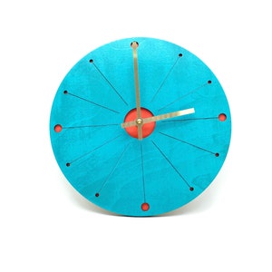 May include: A turquoise blue round clock with a red center circle and gold hands. The clock has a unique design with thin lines radiating from the center.