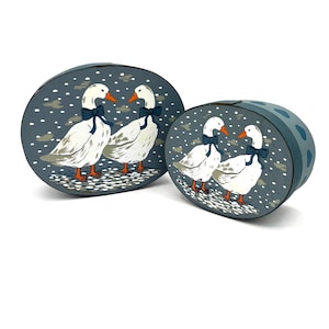 Peut inclure: Deux boîtes décoratives ovales avec un motif d'oies. Les boîtes sont peintes avec un fond bleu foncé et des oies blanches portant des nœuds papillon bleus. Les oies sont dans une scène enneigée. Les boîtes sont de tailles différentes.