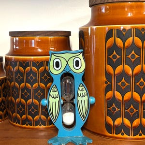 Puede incluir: Un reloj de arena con forma de búho en azul y verde claro con arena blanca, colocado frente a dos grandes recipientes cilíndricos de cerámica color ámbar con un patrón geométrico marrón oscuro y tapas de madera oscura.