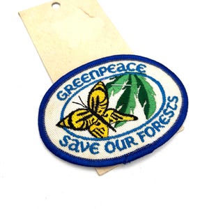 May include: An oval-shaped patch with a blue border and the words "Greenpeace Save Our Forests." The patch features a yellow and black butterfly and green leaves. A tan tag is attached to the patch.