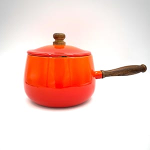 May include: A bright orange enameled metal saucepan with a matching lid. The lid features a small, round, dark brown wooden knob. A long, dark brown wooden handle is attached to the side of the pot, providing a contrast to the vibrant orange finish.