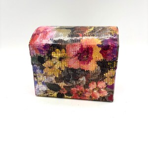 May include: A rectangular box with a rounded top lid featuring a vibrant floral pattern in shades of pink, yellow, orange, and purple. The surface has a textured grid overlay that creates a subtle crosshatch effect over the colourful flower print.