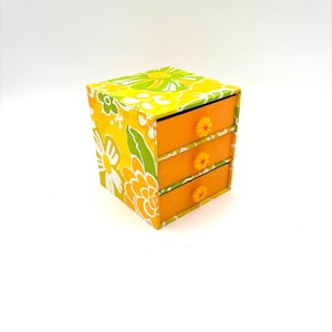 May include: A small, three-drawer storage box with a floral design. The box has a yellow and green floral pattern on the exterior, with orange drawers and flower-shaped knobs. The box is square and sits on a white background.