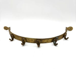 May include: A curved, antique brass wall hook with five hooks. The brass has a weathered appearance, with signs of age and use. The ends of the hook have decorative circular cutouts for mounting. A vintage home decor item.