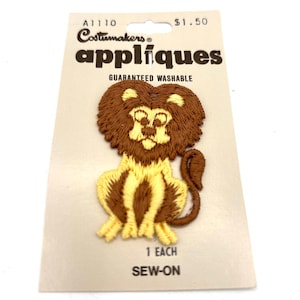 May include: A Costumakers applique of a lion in brown and yellow. The lion is embroidered with a detailed mane, face, and body. The applique is on a cream-colored card with the words "appliqués" and "guaranteed washable".