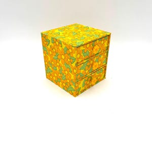 May include: A small, square storage box with two drawers. The box is covered in a bright yellow floral pattern with orange flowers and green leaves. The box is a cube shape, with the drawers visible on the front.