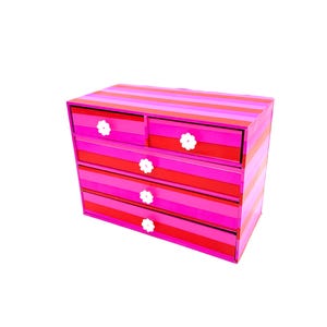 May include: A small, striped chest of drawers in shades of pink and red, with white flower-shaped drawer pulls. The chest has four drawers and a top section with two smaller drawers. The stripes run horizontally across the front and sides.