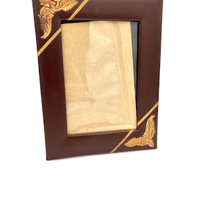 May include: A rectangular dark brown photo frame with a smooth finish. The frame features gold-coloured diagonal lines across the corners and ornate, embossed gold-coloured leaf-like patterns in the top-left and bottom-right corners.