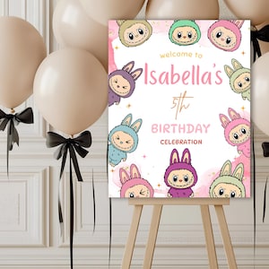 May include: A birthday celebration sign on a wooden easel. The sign is pink and features cartoon bunny characters and the text "Welcome to Isabella's 5th Birthday Celebration". Beige balloons with black bows are in the background.