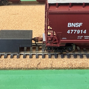 May include: A model train scene featuring a dark red BNSF freight car with the number 477914. A black rectangular structure is connected to the car. The train sits on a model railroad track.