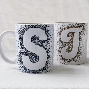 May include: Two white ceramic mugs with a dotted design. One mug has a blue and black dotted 'S' and the other has a brown dotted 'J'.