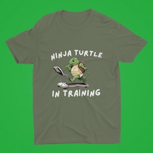 Op de afbeelding: Olijfgroen t-shirt met een cartoon ninja schildpad die op een loopband rent. De witte tekst luidt "NINJA TURTLE IN TRAINING" boven en onder de schildpad-afbeelding.