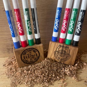 May include: Two wooden pen holders with engraved logos. One holder has the United States Navy logo and the other has a graduate logo with a target. Both holders have five Expo dry erase markers in them: blue, red, green, black, and white.