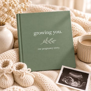 May include: A sage green hardcover book with the white text "growing you." and "our pregnancy story." printed on the front cover, featuring a simple white line drawing of a leafy branch centered between the two lines of text.