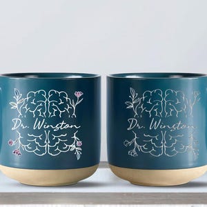 May include: Two teal ceramic mugs with a light tan base. Each mug features a white brain illustration with floral accents and the text "Dr. Winston". The mugs have a curved handle.