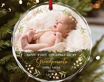 May include: A clear, round Christmas ornament with a baby photo inside. The ornament has a red ribbon for hanging. Text reads "Happy First Christmas as my Grandparents, Love, Lucas 2025" with floral accents. The baby is wrapped in a white blanket.