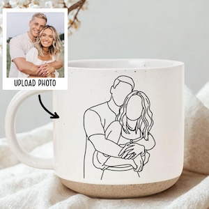 May include: A white ceramic mug with a line drawing of a couple embracing, with a photo and the words "UPLOAD PHOTO" in the upper left corner. The mug has a speckled finish and a tan base. The handle is white.