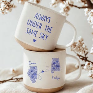 May include: Two white ceramic mugs stacked on top of each other. The top mug has the text "ALWAYS UNDER THE SAME SKY" in blue. The bottom mug features state outlines with floral designs, names, and a connecting airplane graphic.