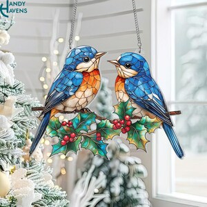 May include: A decorative hanging ornament featuring two bluebirds perched on a branch with holly and red berries. The birds have orange chests and are set against a window with a Christmas tree in the background. The text "ANDY HAVENS" is in the upper left corner.