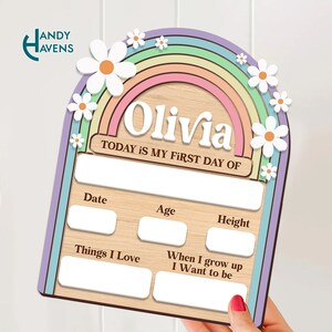 May include: A wooden growth chart with a rainbow and daisies. The chart has spaces to fill in the child's name, date, age, height, things they love, and what they want to be when they grow up. The text "TODAY IS MY FIRST DAY OF" is printed on the chart. The chart is light brown with a purple, green, blue, pink, and orange rainbow.