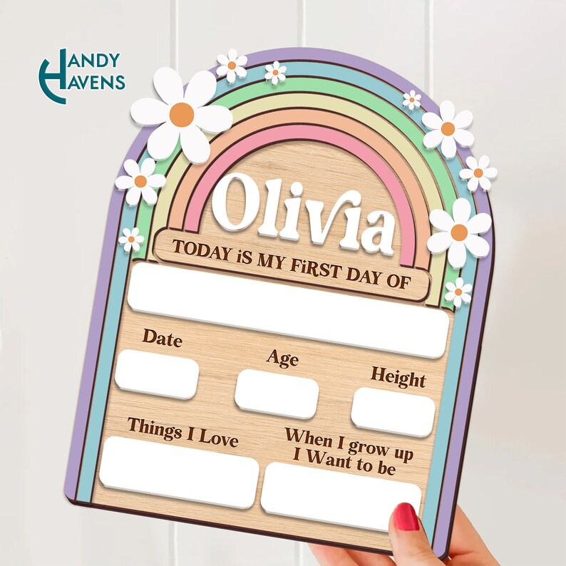 May include: A wooden growth chart with a rainbow and daisies. The chart has spaces to fill in the child's name, date, age, height, things they love, and what they want to be when they grow up. The text "TODAY IS MY FIRST DAY OF" is printed on the chart. The chart is light brown with a purple, green, blue, pink, and orange rainbow.