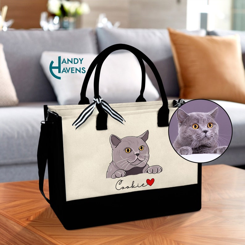 K&ouml;nnte beinhalten: Eine schwarz-wei&szlig;e Tragetasche mit einem schwarzen Griff und einer wei&szlig;en Canvas-Vorderseite. Die Vorderseite der Tasche zeigt eine Cartoon-Illustration einer grauen Katze mit dem Namen "Cookie" darunter.