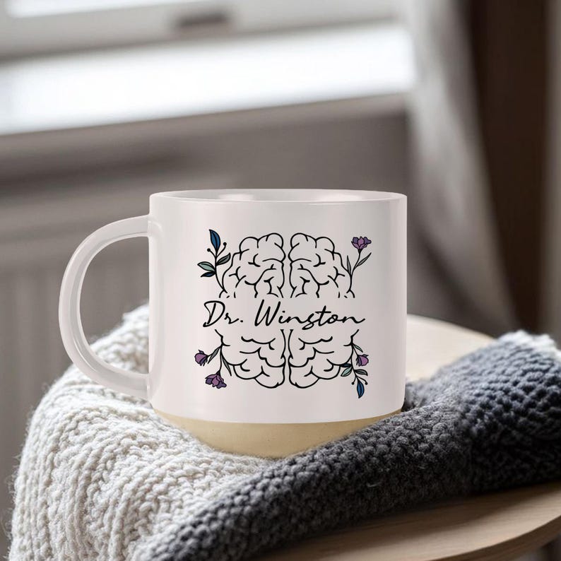 May include: White ceramic mug with a cream-colored base, featuring a black brain illustration and the name "Dr. Winston" in script. The design includes floral accents in purple and green. The mug is on a wooden surface.