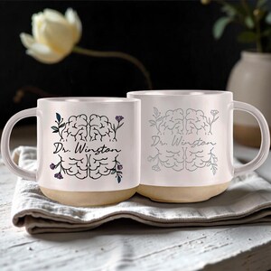May include: Two white ceramic mugs with a tan base. Each mug features a brain illustration with floral accents and the text "Dr. Winston". The mugs are on a white cloth, with a blurred flower in the background.