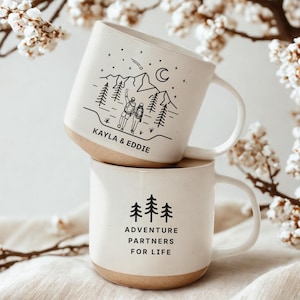 May include: Two white ceramic mugs stacked on top of each other. The top mug has a black illustration of a mountain scene with two people and the names "KAYLA & EDDIE". The bottom mug has the text "ADVENTURE PARTNERS FOR LIFE" in black.