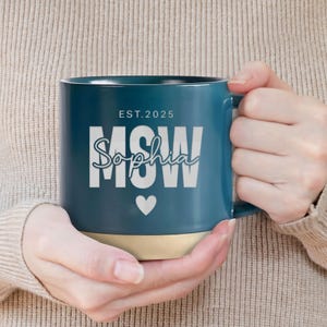 May include: A teal blue ceramic mug with a white inscription that reads "EST. 2025 MSW Sophia" with a white heart symbol below the text. The mug has a beige rim.