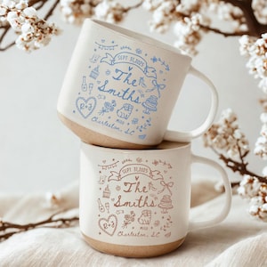 May include: Two stacked white ceramic mugs with a light brown base. Each mug has a hand-drawn design in blue and pink, including the text "The Smiths," "Charleston, SC," and the date "SEPT 31, 2025." The mugs have a handle and are decorated with hearts, flowers, and a cake.
