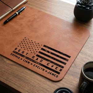 May include: A brown leather desk mat with the text "BEST DAD EVER" and an American flag design. The names Sophia, Liam, Evelyn, and James are also included. A black pen and notebook are visible.