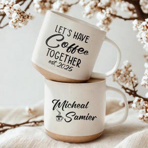 May include: Two stacked white ceramic coffee mugs with brown bases. The top mug has the text "LET'S HAVE Coffee TOGETHER est. 2025" in black script. The bottom mug displays the name "Micheal Samier" in black script with a coffee cup icon.