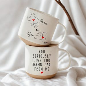May include: Two white ceramic mugs stacked on top of each other. The top mug features a map of Texas and New York with names and a dotted line with an airplane. The bottom mug has the text "YOU SERIOUSLY LIVE TOO DAMN FAR FROM ME" with a red heart.