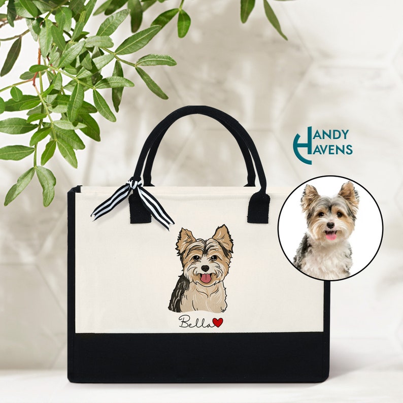 K&ouml;nnte beinhalten: Eine wei&szlig;e Canvas-Tragetasche mit schwarzen Henkeln und einem schwarzen Boden. Die Tasche hat ein schwarz-wei&szlig;es Band, das um die Henkel gebunden ist. Die Tasche zeigt ein Cartoon-Bild eines Yorkshire Terriers mit dem Namen "Bella" und einem roten Herzen.