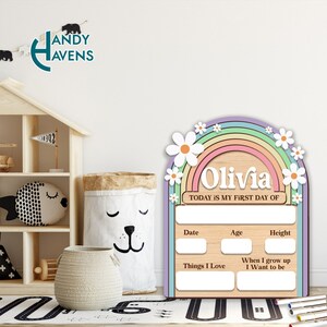 May include: A wooden growth chart with a rainbow and daisies design. The chart has spaces to write the child's name, date, age, height, things they love, and what they want to be when they grow up. The text "Olivia" and "TODAY IS MY FIRST DAY OF" are written on the chart.