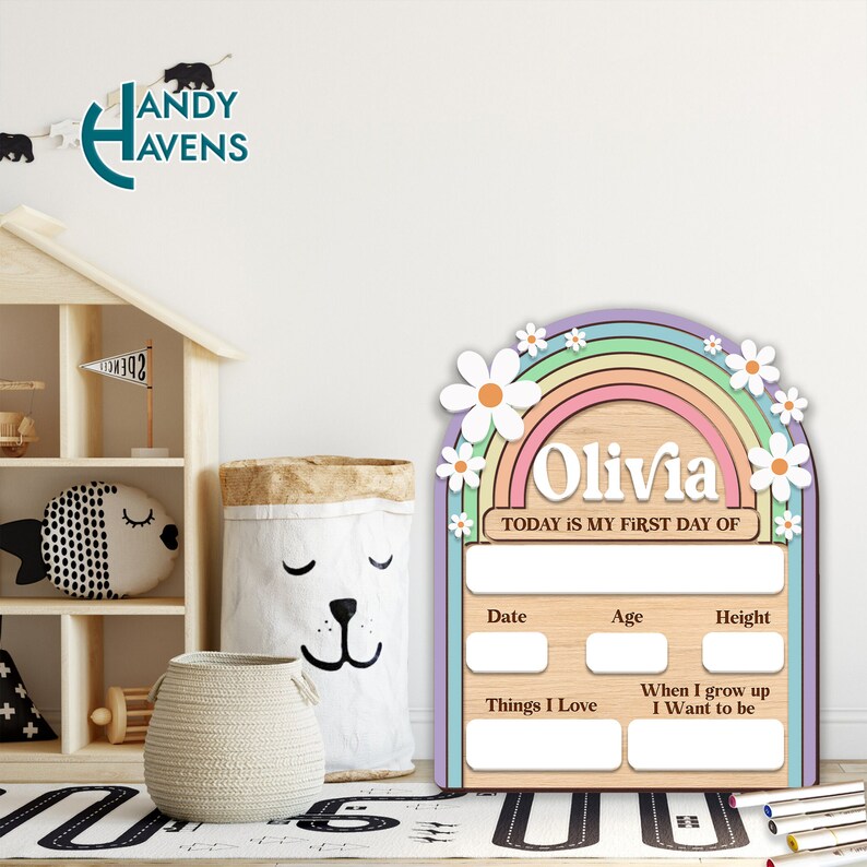 May include: A wooden growth chart with a rainbow and daisies design. The chart has spaces to write the child's name, date, age, height, things they love, and what they want to be when they grow up. The text "Olivia" and "TODAY IS MY FIRST DAY OF" are written on the chart.