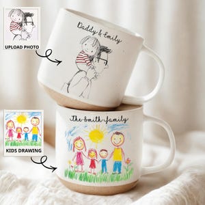 May include: Two white ceramic mugs stacked on top of each other. The top mug has a drawing of a person carrying a child on their shoulders, with the text "Daddy & Emily". The bottom mug has a drawing of a family with the text "The Smith family".