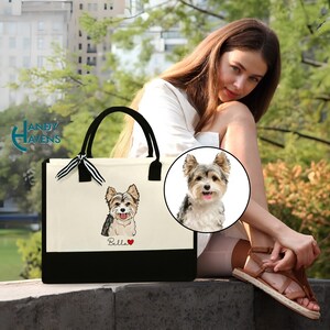 K&ouml;nnte beinhalten: Eine wei&szlig;e Canvas-Tragetasche mit schwarzen Henkeln und einem schwarzen Boden. Die Tasche zeigt eine Schwarzwei&szlig;-Illustration eines Yorkshire Terriers mit dem Namen "Bella" unter dem Hund.