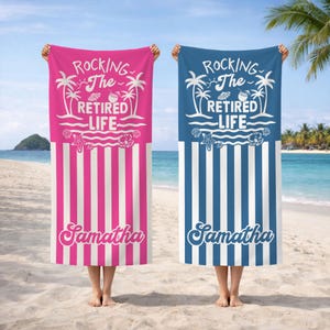 May include: Two beach towels, one pink and one blue, are displayed on a sandy beach. The towels feature white vertical stripes at the bottom and the text "Rocking The Retired Life" with a beach scene graphic. Each towel is personalised with the name "Samantha".