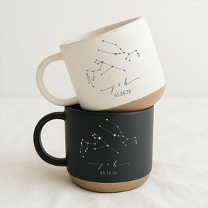 May include: Two ceramic mugs, one cream-colored and one black, stacked on top of each other. Each mug features a constellation design and the inscription "j + h 02.28.26". The mugs have a natural, unglazed base and a curved handle.