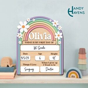 May include: A wooden sign with a rainbow and daisies, personalized with the name "Olivia". It says "Today is my first day of 1st Grade" and includes details like date, age, height, things they love, and what they want to be when they grow up.
