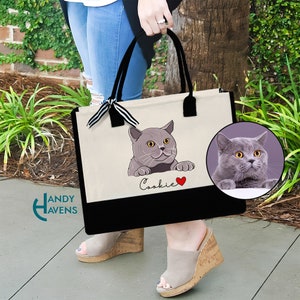 K&ouml;nnte beinhalten: Eine wei&szlig;e Canvas-Tragetasche mit schwarzen Henkeln und einem schwarzen Boden. Die Tasche zeigt ein Cartoon-Bild einer grauen Katze mit dem Namen "Cookie" darunter. Ein rotes Herz befindet sich neben dem Namen. Ein Nahaufnahmebild der Katze ist in der oberen rechten Ecke des Bildes zu sehen.