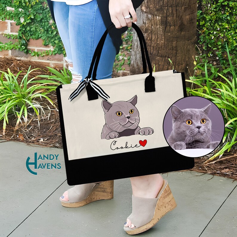K&ouml;nnte beinhalten: Eine wei&szlig;e Canvas-Tragetasche mit schwarzen Henkeln und einem schwarzen Boden. Die Tasche zeigt ein Cartoon-Bild einer grauen Katze mit dem Namen "Cookie" darunter. Ein rotes Herz befindet sich neben dem Namen. Ein Nahaufnahmebild der Katze ist in der oberen rechten Ecke des Bildes zu sehen.
