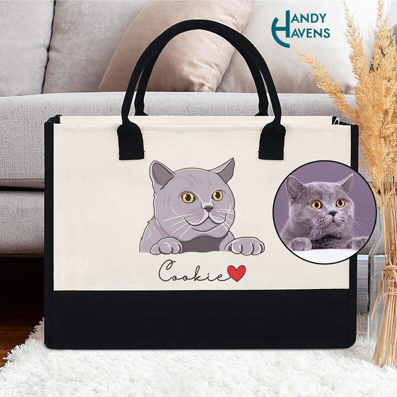 K&ouml;nnte beinhalten: Eine wei&szlig;e Canvas-Tragetasche mit schwarzen Henkeln und einem schwarzen Boden. Die Tasche zeigt eine Cartoon-Illustration einer grauen Katze mit gelben Augen und dem Text "Cookie" mit einem roten Herzen.