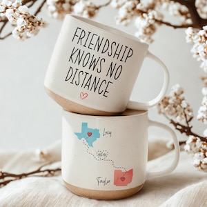 May include: Two white ceramic mugs stacked on top of each other. The top mug has the text "FRIENDSHIP KNOWS NO DISTANCE" in black, with a pink heart. The bottom mug features a map design with the names "Lucy" and "Taylor".